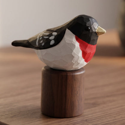 A geometric, faceted wooden Rose-breasted Grosbeak figurine with a black, white, and red painted design, perched on a cylindrical dark brown wooden base, set on a wooden tabletop.