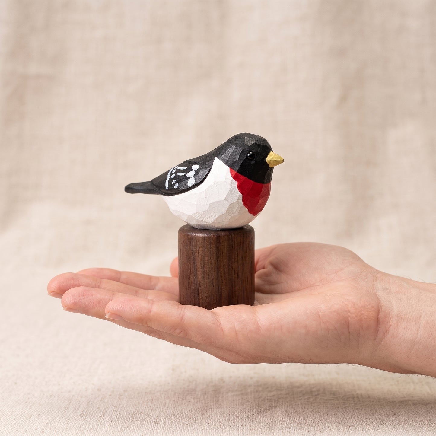 A small carved Rose-breasted Grosbeak figurine with a black, white, and red body and a yellow beak, perched on a dark wooden cylindrical base and held in a person's hand against a soft beige fabric background.