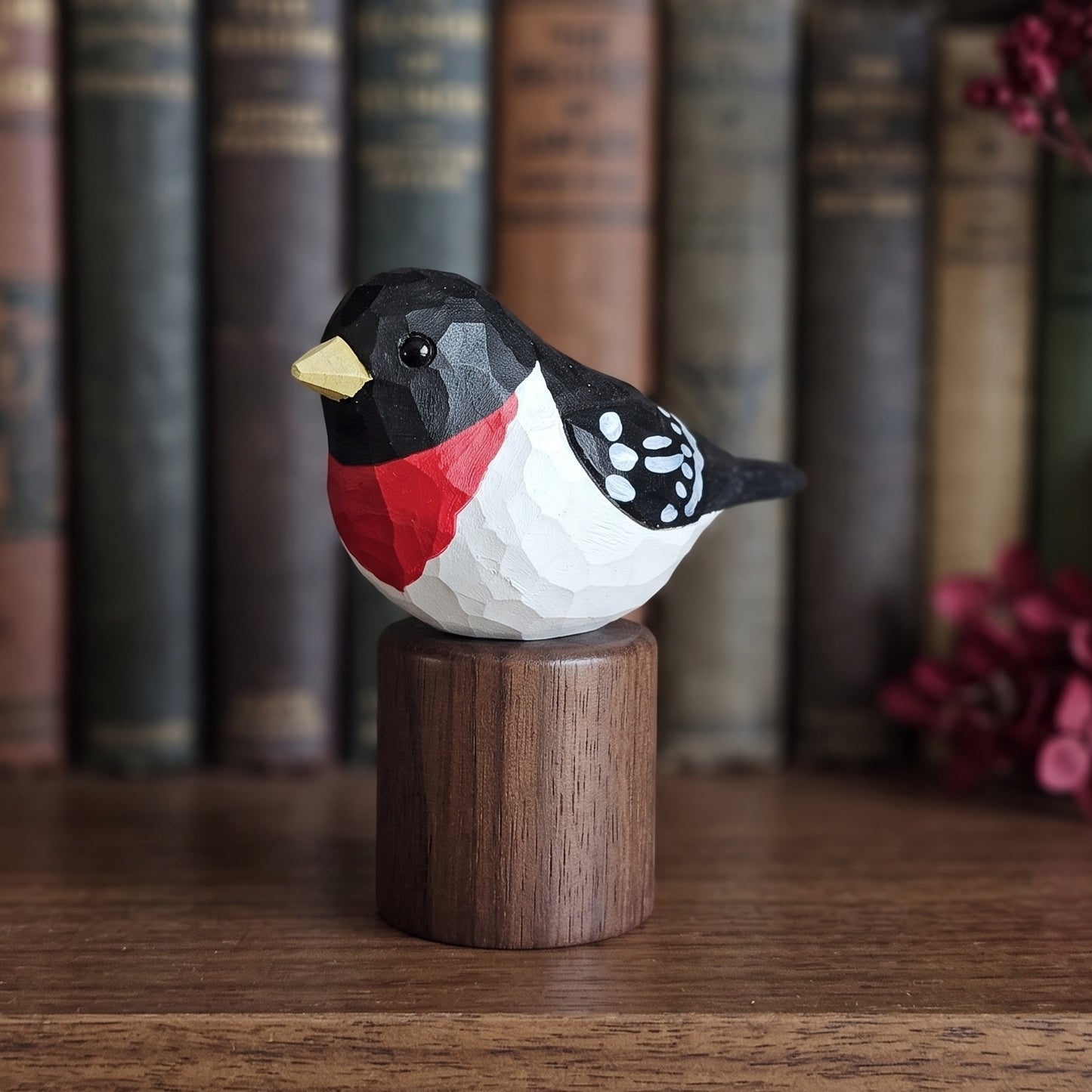 A low-poly wooden Rose-breasted Grosbeak figurine with a red chest, black and white body, and yellow beak, mounted on a round wooden base on a wooden table with a blurred background of books.