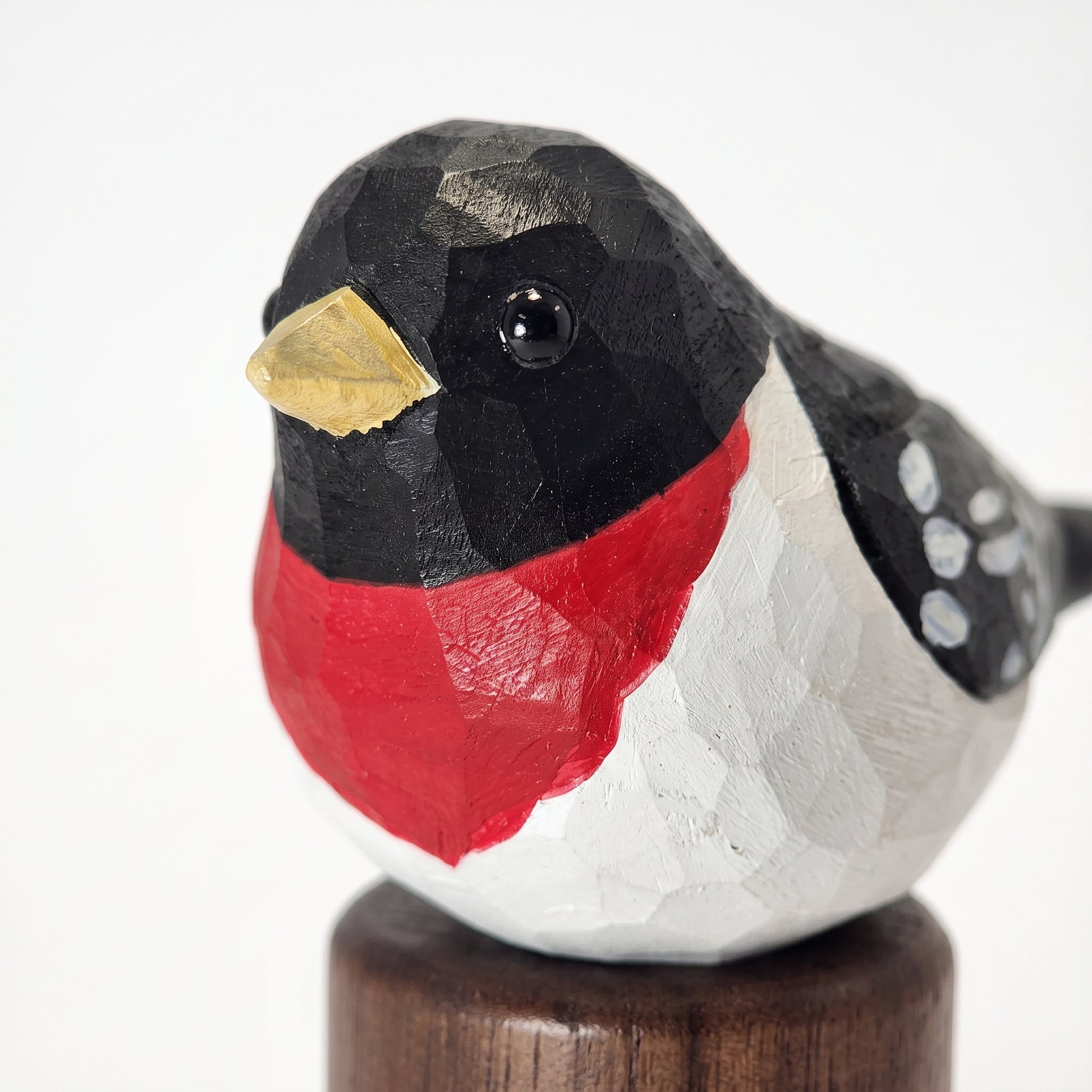 A hand-carved wooden Rose-breasted Grosbeak figurine with a faceted geometric texture, painted in a black, white, and red bird-of-prey style with a gold beak, mounted on a dark wooden base against a white background.