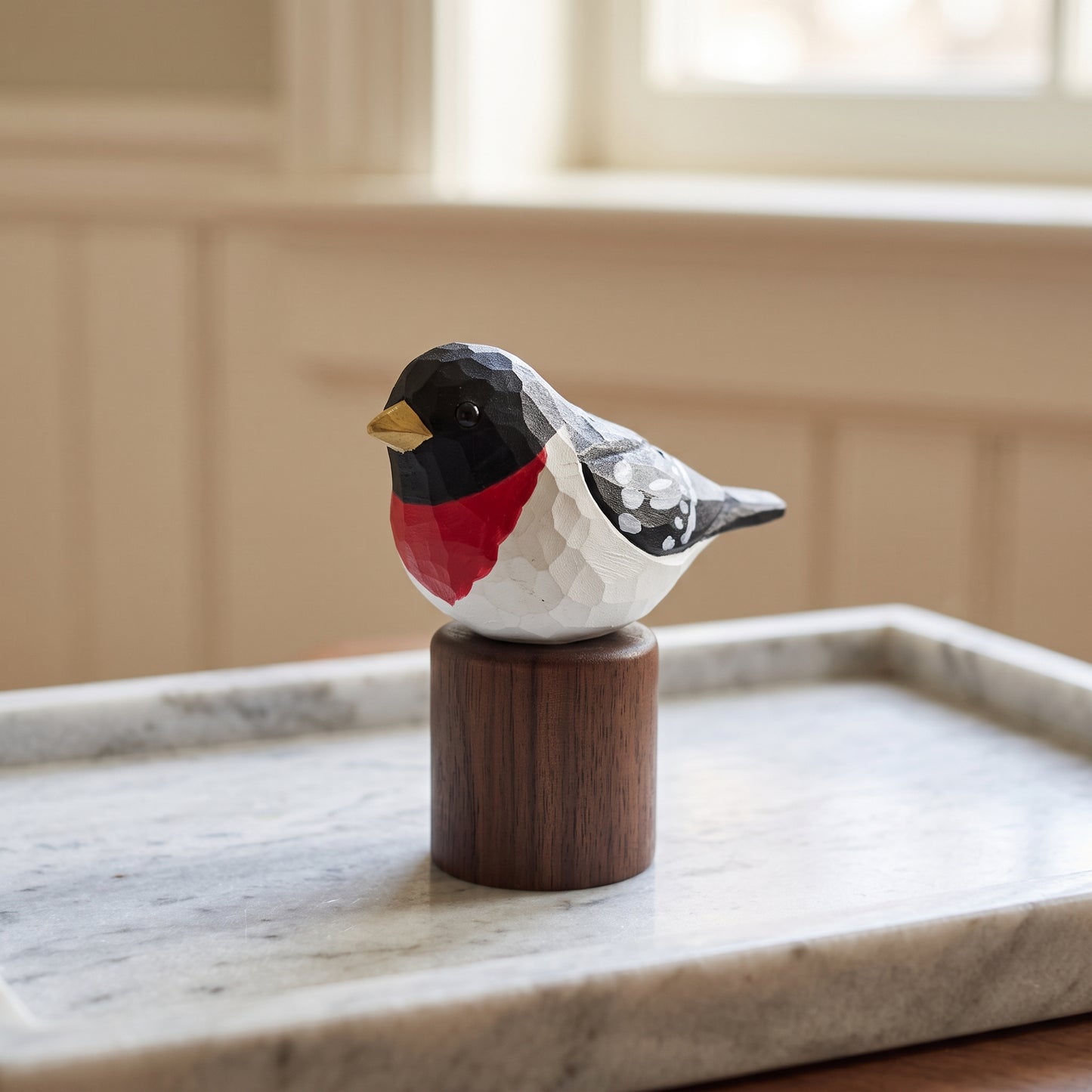 A hand-carved wooden Rose-breasted Grosbeak  figurine with a red, white, and black patterned body and a yellow beak, perched on a dark wood cylinder base. The figurine is placed on a light marble tray in a softly blurred indoor setting.