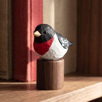 A geometric, faceted Rose-breasted Grosbeak figurine with a black, white, and red pattern and a gold beak, perched on a small cylindrical wooden base on a wooden shelf.