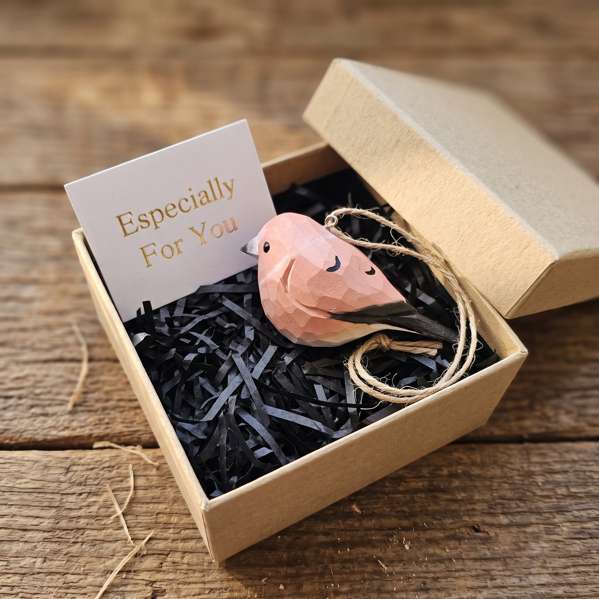A small geometric pink Rosefinch ornament with black accents and twine, presented in a kraft gift box with shredded black filler and a card that reads 'Especially For You' on a rustic wooden surface.