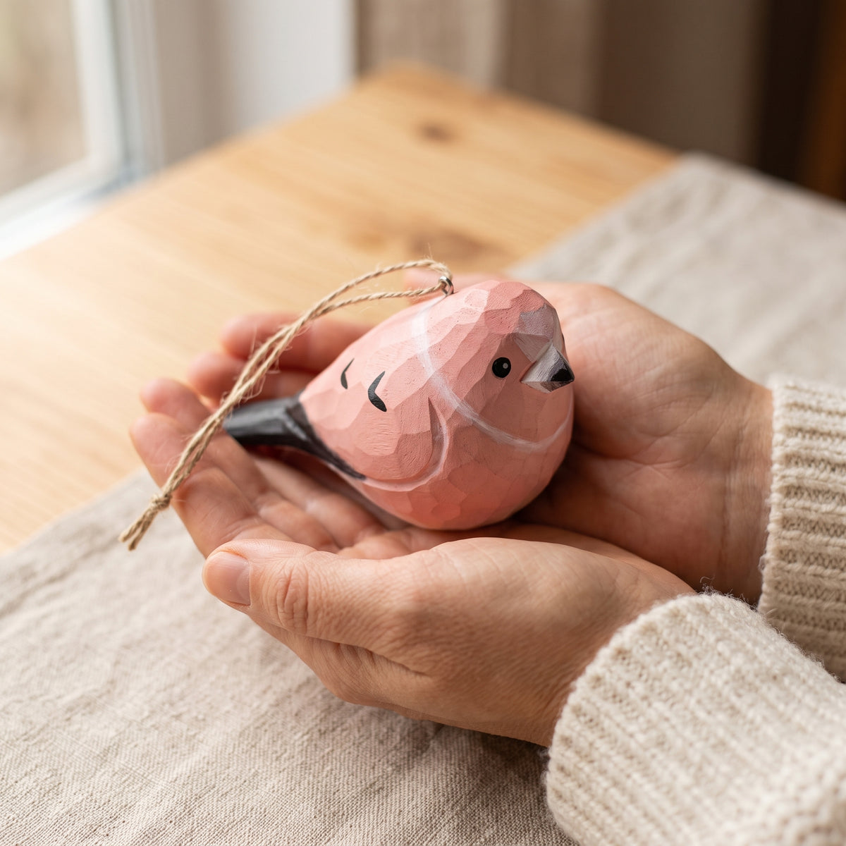 A small faceted wooden Rosefinch ornament in soft pink with black and white details, held gently in two hands over a beige knit sweater, with a wooden table and softly blurred indoor background.
