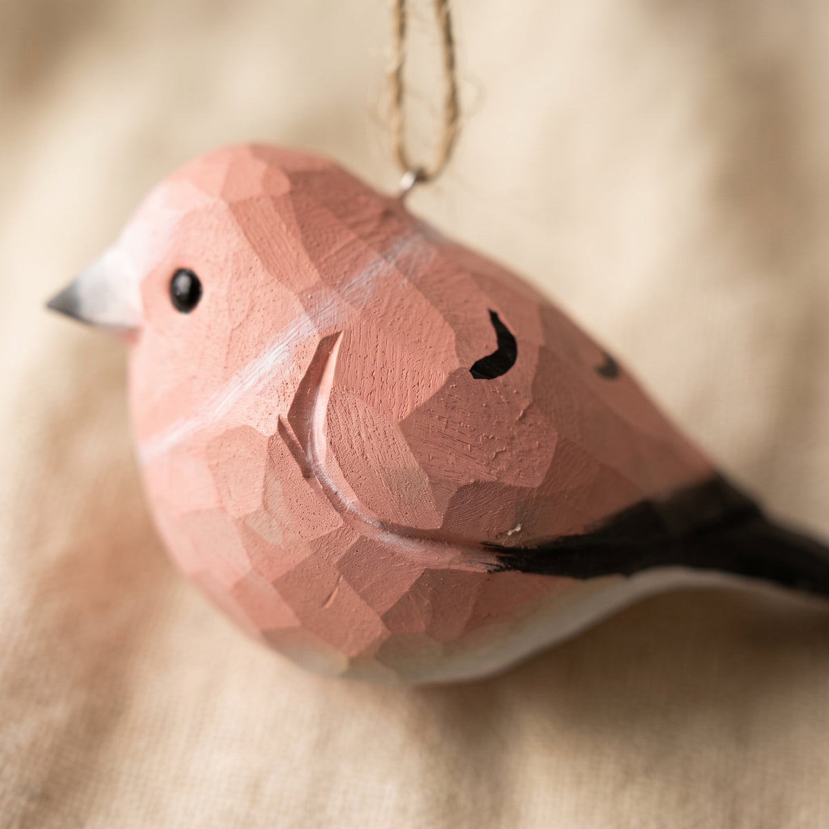 A close-up of a small Rosefinch-shaped hanging ornament with a faceted geometric texture. The ornament is primarily a dusty pink color with a black beak, black eye, black curved line detail, and black tail feathers.