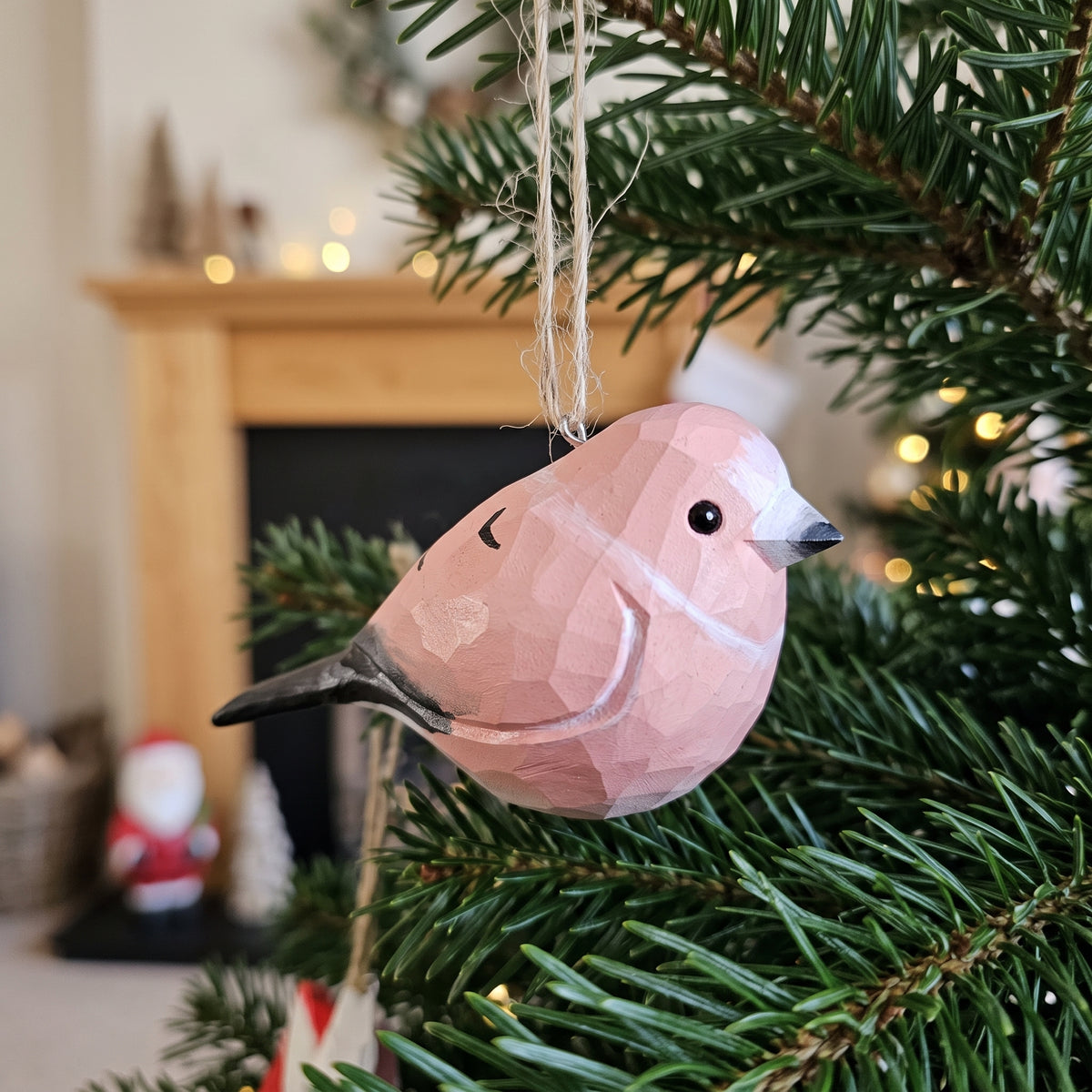 A pink faceted Rosefinch Christmas ornament hanging from a string on a green Christmas tree in front of a softly blurred fireplace background.