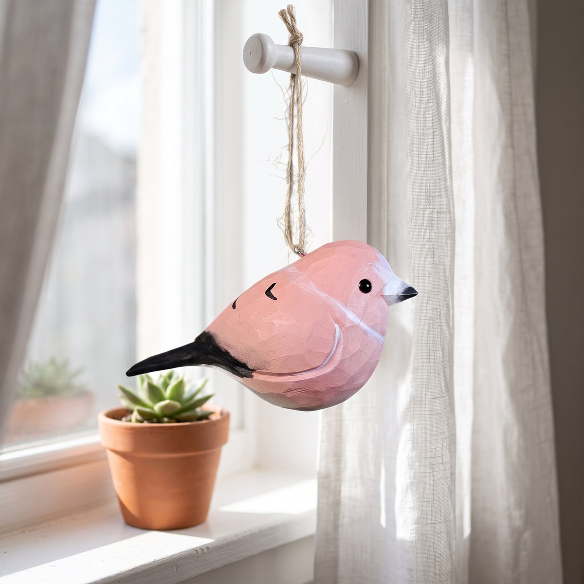 A pink wooden Rosefinch hanging ornament with a black beak and tail, suspended by a jute string from a white window rod in a sunlit room with sheer curtains and a small potted succulent on the sill.