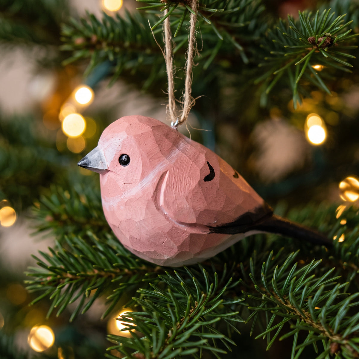 A pink faceted Rosefinch Christmas ornament hanging from a jute string on a green pine tree with warm bokeh lights in the background.