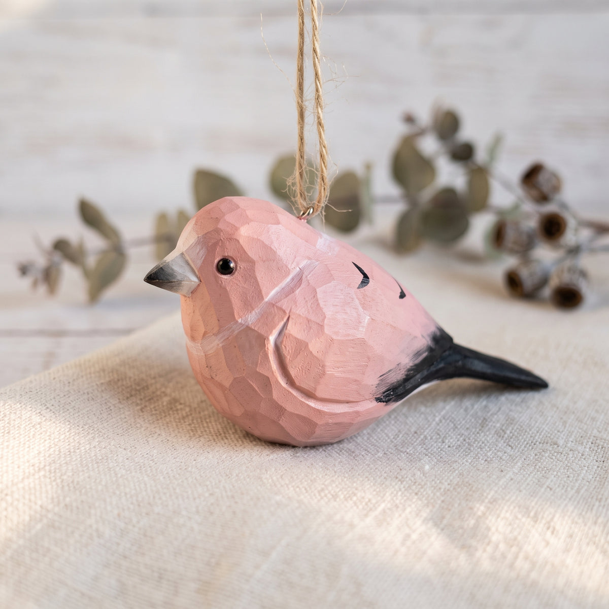 A small carved wooden Rosefinch ornament in a dusty pink color with a black tail and beak, hanging from a natural jute string against a rustic, light beige background with blurred eucalyptus branches.