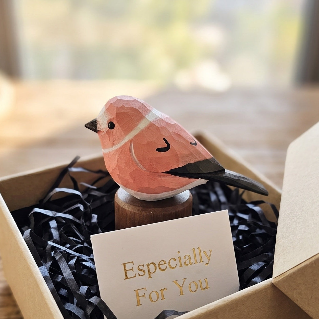 A small pink Rosefinch figurine perched on a wooden stand, displayed inside an open kraft gift box with black shredded paper and a card that reads 'Especially For You.' The box sits on a wooden table in a softly blurred indoor setting.