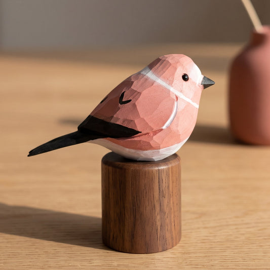 A geometric Rosefinch figurine with a pink, peach, and white faceted body, black wings and beak, perched on a small round wooden base. The figurine is photographed on a light wood tabletop with a blurred terracotta vase and dried flower in the background.