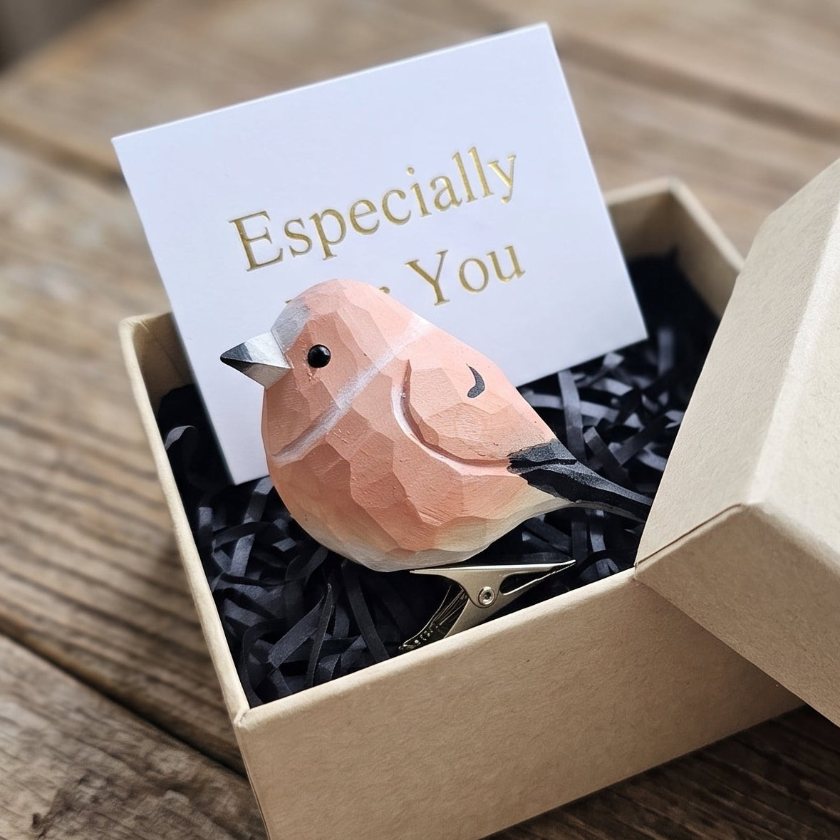 A hand-carved wooden Common Rosefinch clip-on bird ornament resting inside an open kraft paper gift box filled with black shredded paper, featuring rose-pink and coral-red painted plumage, a small dark eye, and subtle carved feather details, alongside a white card reading "Especially For You" placed in the foreground on a rustic wooden surface.