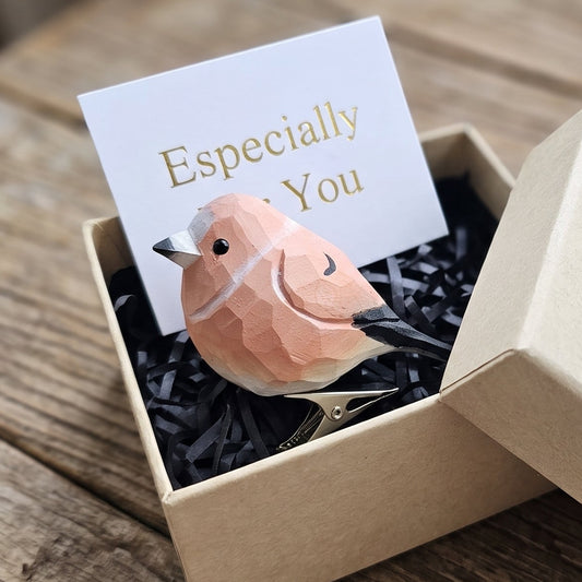 A hand-carved wooden Common Rosefinch clip-on bird ornament resting inside an open kraft paper gift box filled with black shredded paper, featuring rose-pink and coral-red painted plumage, a small dark eye, and subtle carved feather details, alongside a white card reading "Especially For You" placed in the foreground on a rustic wooden surface.