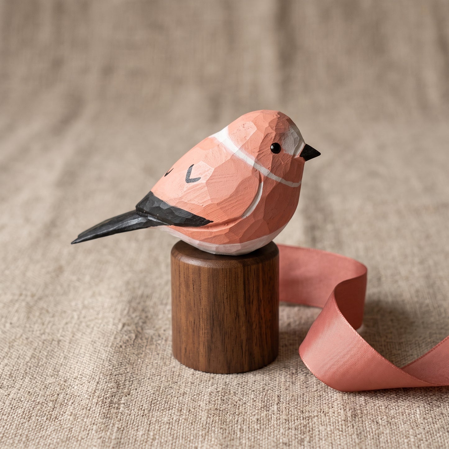 A low-poly style wooden Rosefinch figurine in terracotta pink and white with black accents, perched on a small cylindrical wooden base with a pink ribbon trailing beside it, set against a textured beige fabric background.