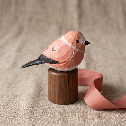 A low-poly style wooden Rosefinch figurine in terracotta pink and white with black accents, perched on a small cylindrical wooden base with a pink ribbon trailing beside it, set against a textured beige fabric background.