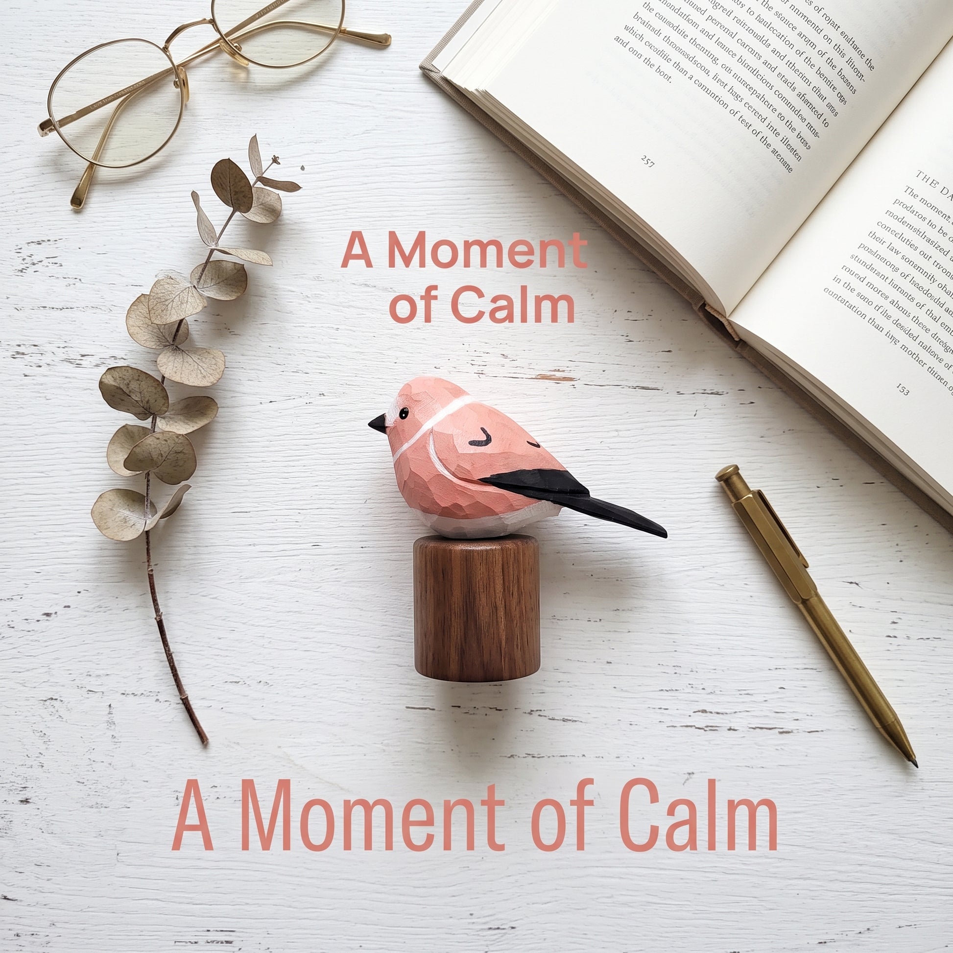 A pink and black Rosefinch figurine on a small wooden block, styled in a flat lay with an open book, eyeglasses, dried eucalyptus leaves, and a gold pen on a white wooden surface. Text overlays read 'A Moment of Calm'.