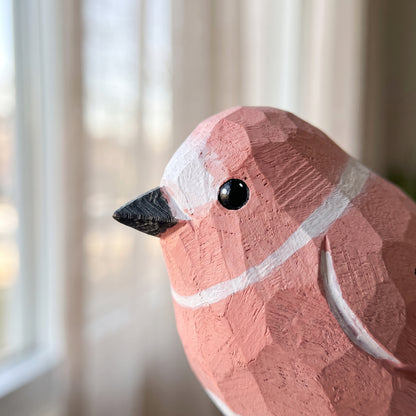 A close-up of a carved wooden Rosefinch figurine with a pinkish-terracotta finish, white painted accents, and a black beak, set against a softly blurred indoor background.