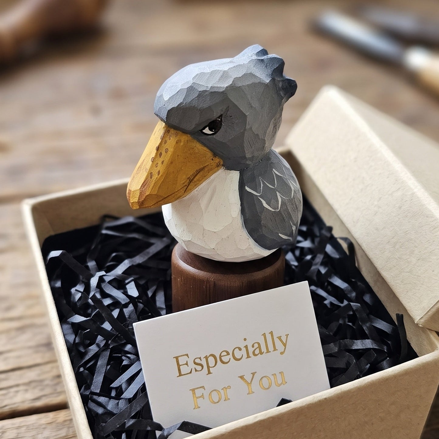 A carved wooden Baby Shoebill figurine with a gray head, white chest, and yellow beak, displayed in a kraft gift box filled with black shredded paper and a small card that reads 'Especially For You'.