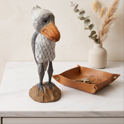 A stylized, low-poly Shoebill figurine with a gray and white patterned body and a brown beak, standing on a rock base next to a brown leather key tray on a white marble surface.