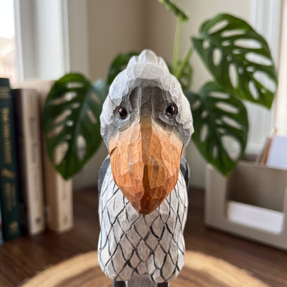 A hand-carved wooden Shoebill figurine, likely a toucan or bearded bird, with a textured gray and white patterned body and a warm orange-brown beak, set on a wooden surface with a blurred background of books and a green plant.