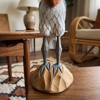 A handcrafted wooden Shoebill figurine with detailed gray and white feathers, standing on a carved natural wood base on a wooden table in a cozy living room setting.