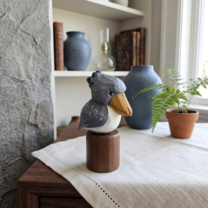 A geometric wooden Baby Shoebill  figurine with a gray, white, and orange-brown body, mounted on a cylindrical wooden base and displayed on a table runner in a styled home interior.