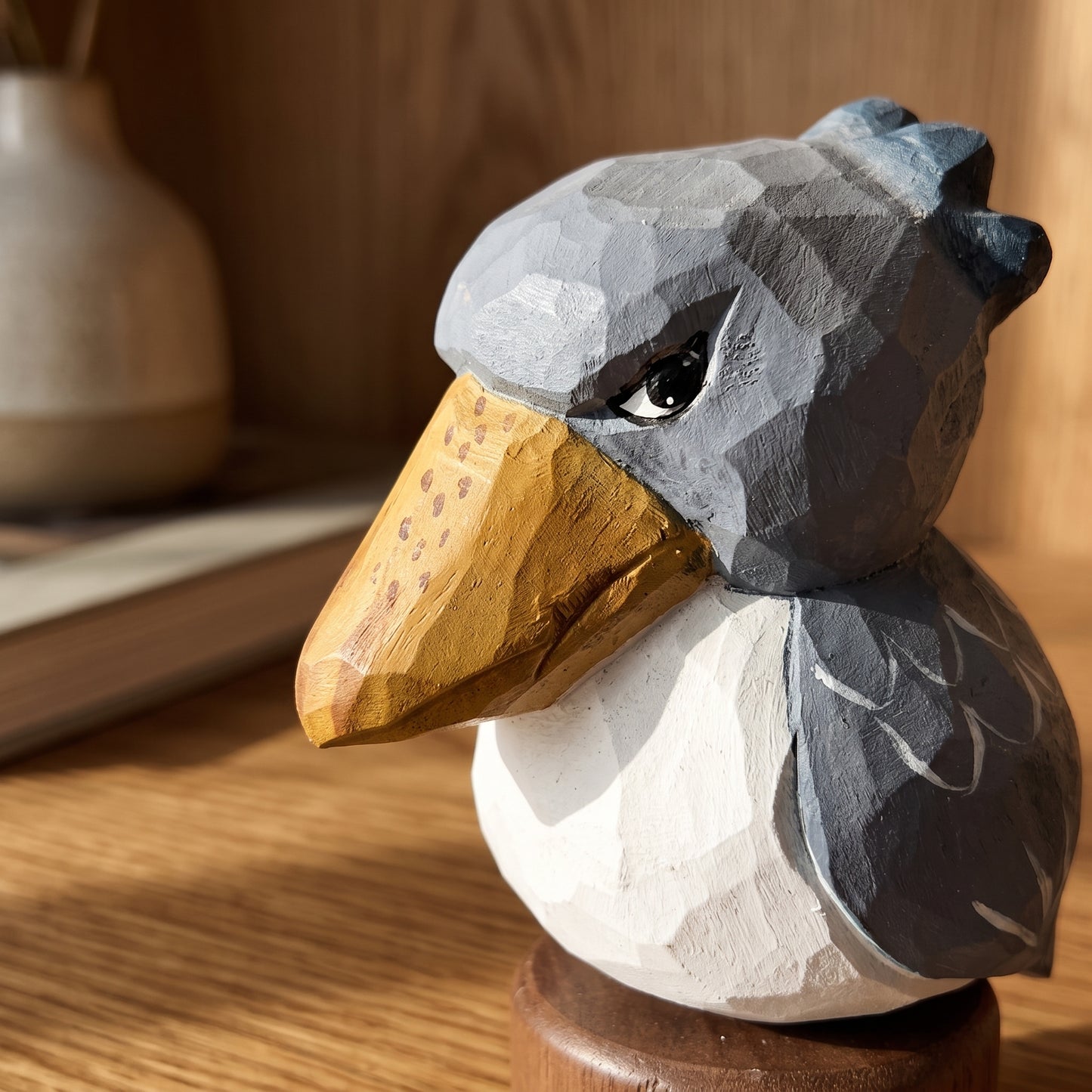 A low-poly wooden Baby Shoebill sculpture with a gray head and wings, a white body, and a carved natural wood beak, displayed on a wooden surface.