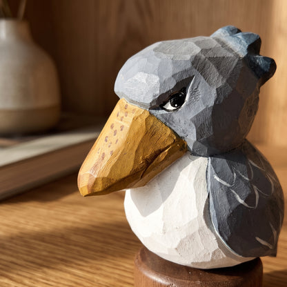A low-poly wooden Baby Shoebill sculpture with a gray head and wings, a white body, and a carved natural wood beak, displayed on a wooden surface.