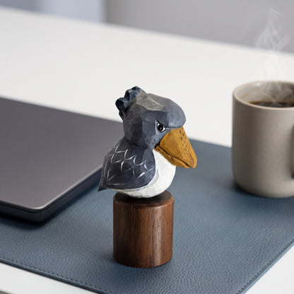 A small sculptural Baby Shoebill figurine with a dark gray head and back, white chest, and mustard-yellow beak, mounted on a round wooden base. It is photographed on a desk with a laptop, a mug of coffee, and a blue mat in the background.