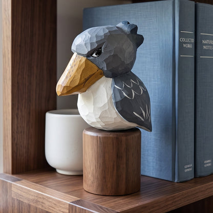 A geometric, faceted Baby Shoebill  sculpture in gray, white, and yellow sits on a wooden pedestal on a dark wood bookshelf, styled between blue books and a white ceramic cup.