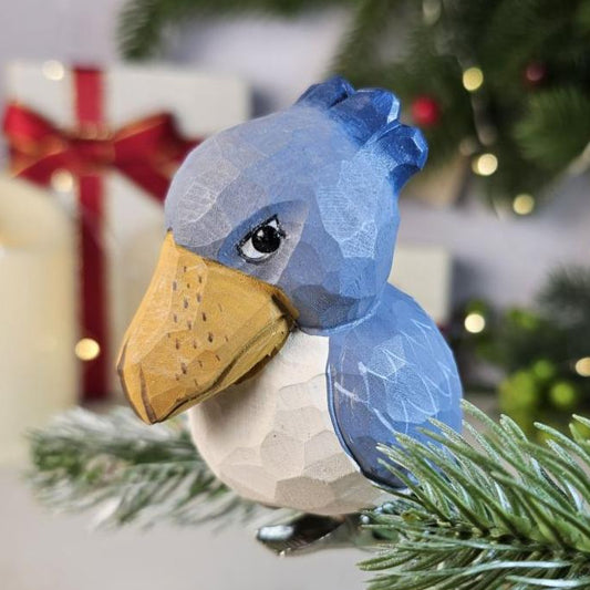A wooden Shoebill clip-on Christmas tree ornament bird, with blue, white, and yellow coloring, attached to a Christmas tree branch.