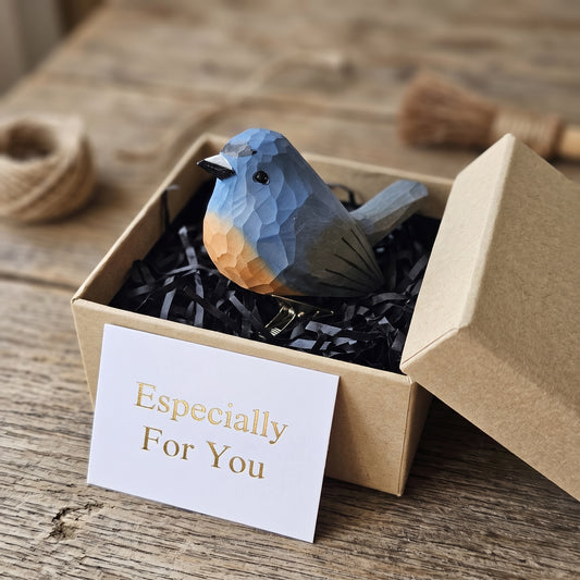 A hand-carved wooden Snowy-Browed Flycatcher ornament displayed inside an open kraft gift box filled with black shredded paper, featuring a light blue-grey body, a soft peach-orange throat and breast, a silver metal clip attached underneath, and a white card reading "Especially For You" placed in the foreground, set against a rustic wooden surface.