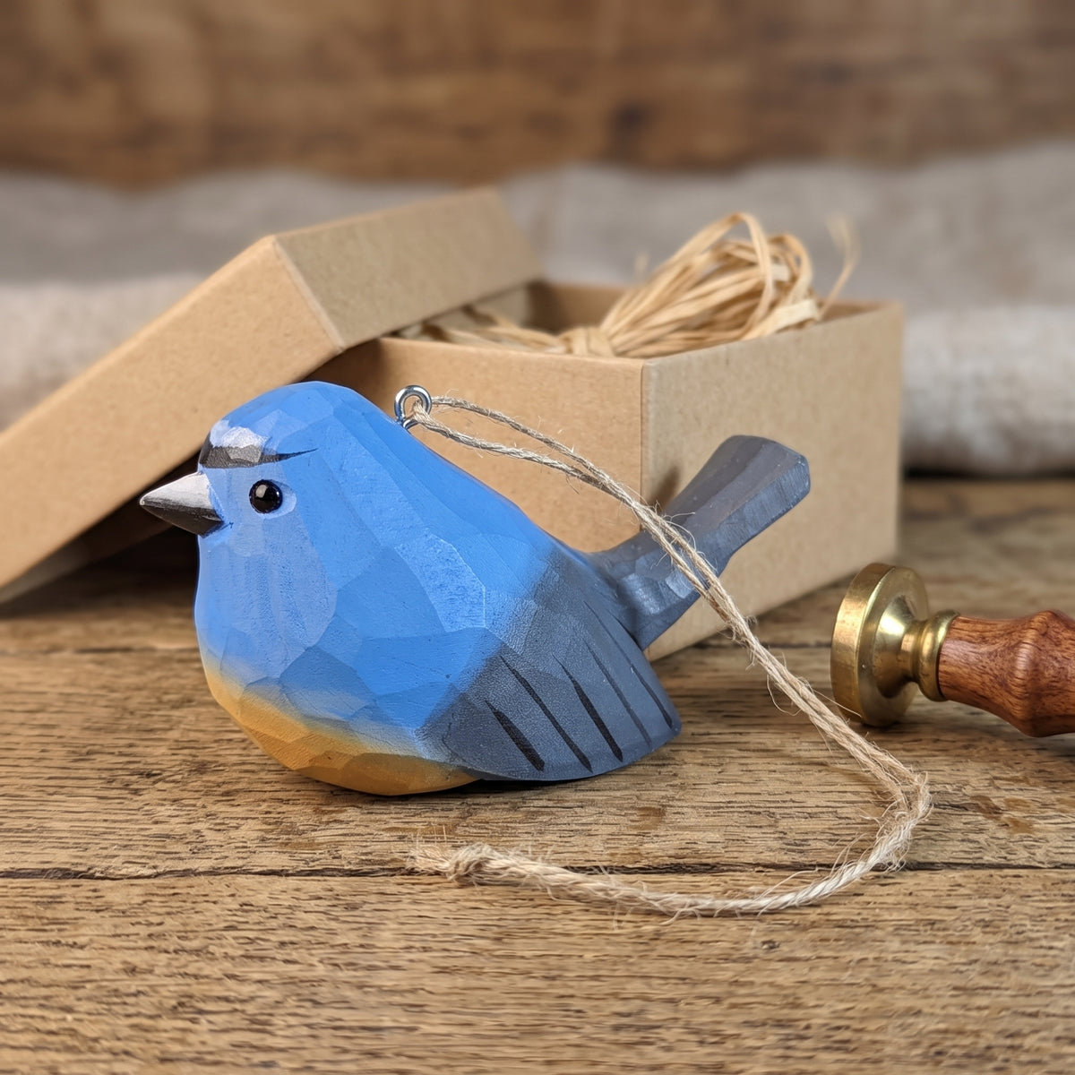 A handcrafted wooden Snowy-browed flycatcher ornament painted in shades of blue, gray, and natural wood tones, with a small metal loop at the top for hanging. It is displayed on a rustic wooden surface next to its open kraft paper gift box.