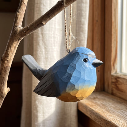 A geometric, faceted wooden Snowy-browed flycatcher ornament in shades of blue, gray, and yellow, hanging from a natural twine loop on a rustic wooden branch in front of a window.