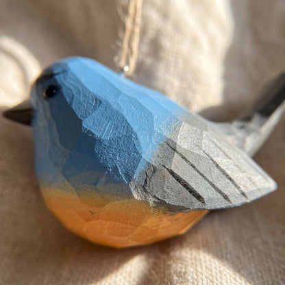 A handcrafted wooden Snowy-browed flycatcher bird ornament with a faceted geometric design, featuring a blue and orange gradient painted finish, resting on a textured beige fabric background.