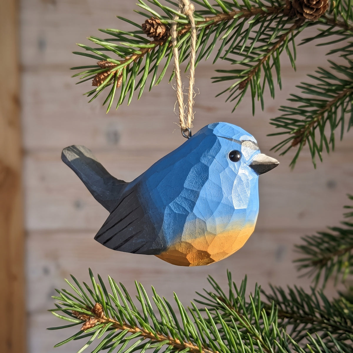 A geometric, faceted Snowy-browed flycatcher ornament in shades of blue and orange hangs from a natural twine string on a pine tree branch, with a soft-focus wood panel background.