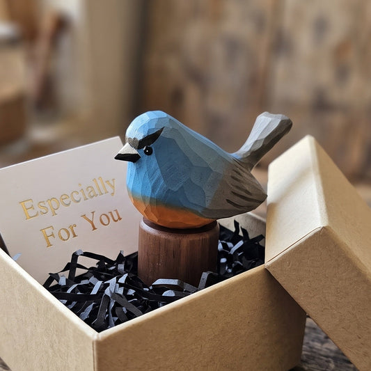 A small wooden Snowy-browed flycatcher figurine with a blue-gray body, orange chest, and black beak, displayed inside an open kraft gift box with shredded black paper filler and a card that reads 'Especially For You.' The box sits on a rustic wooden table with blurred tools and wood in the background.