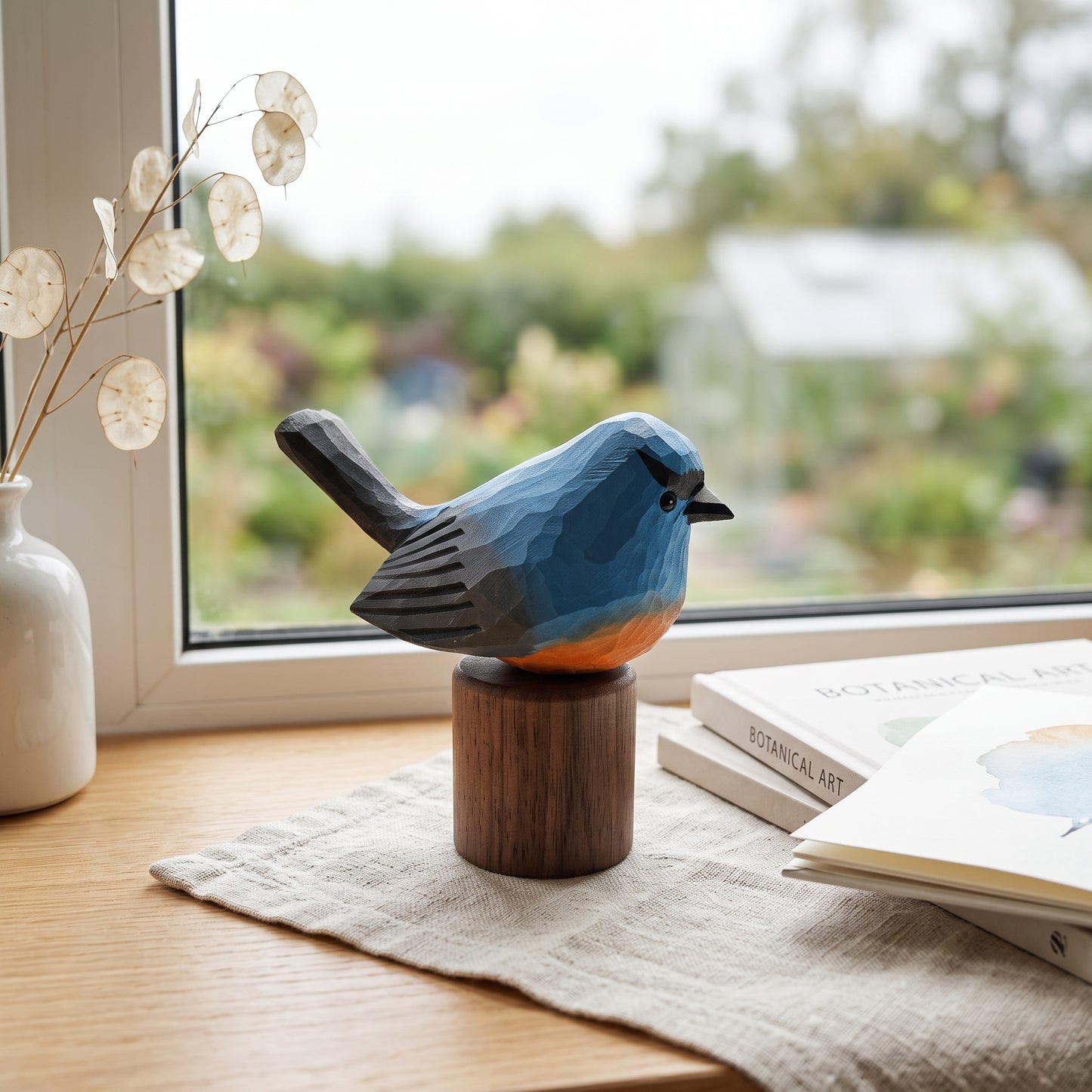 A geometric, faceted wooden Snowy-browed flycatcher figurine in blue, orange, and black, mounted on a round wooden pedestal. The figurine is styled on a linen runner on a wooden table next to a window with a garden view, with a white vase and books in the background.