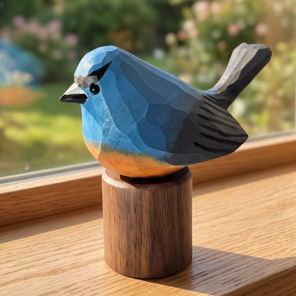 A geometric wooden Snowy-browed flycatcher sculpture in shades of blue, gray, and orange, mounted on a cylindrical wooden base and placed on a wooden windowsill with a blurred garden view in the background.