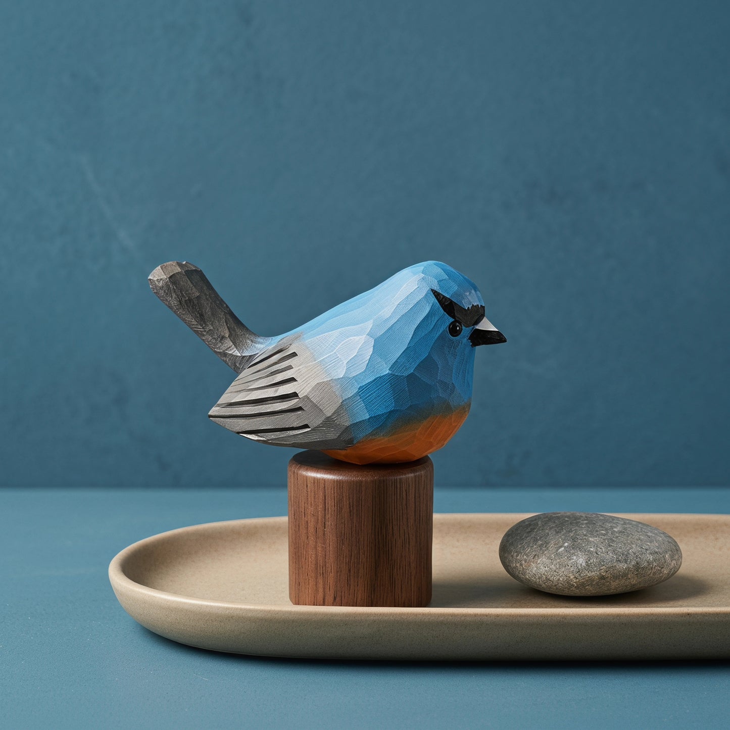 A geometric, faceted wooden Snowy-browed flycatcher sculpture in shades of blue, gray, and orange, mounted on a cylindrical wooden pedestal. The sculpture is styled on a beige ceramic tray next to a smooth gray stone against a textured blue background.