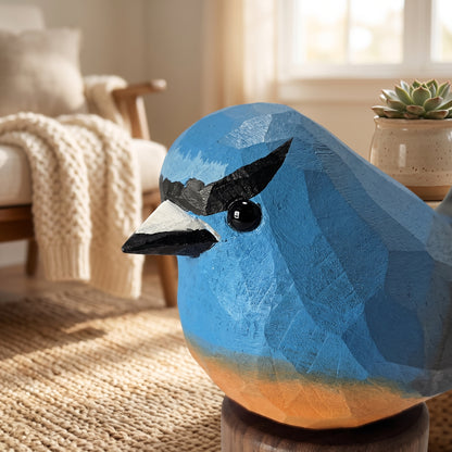 A geometric, faceted Snowy-browed flycatcher sculpture in shades of blue with a black beak and eye, displayed on a wooden stand in a cozy, softly lit living room.