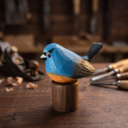 A hand-carved wooden Snowy-browed flycatcher figurine with a bright blue back, black beak and tail, and a warm orange belly, mounted on a small cylindrical wooden base on a dark workbench.