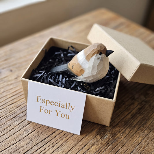 A hand-carved wooden House Sparrow clip-on ornament resting inside an open kraft gift box with black shredded paper filling, featuring a light brown head with a grey crown and white cheeks, a silver metal clip underneath, and a white card reading "Especially For You" placed in the foreground on a wooden tabletop.