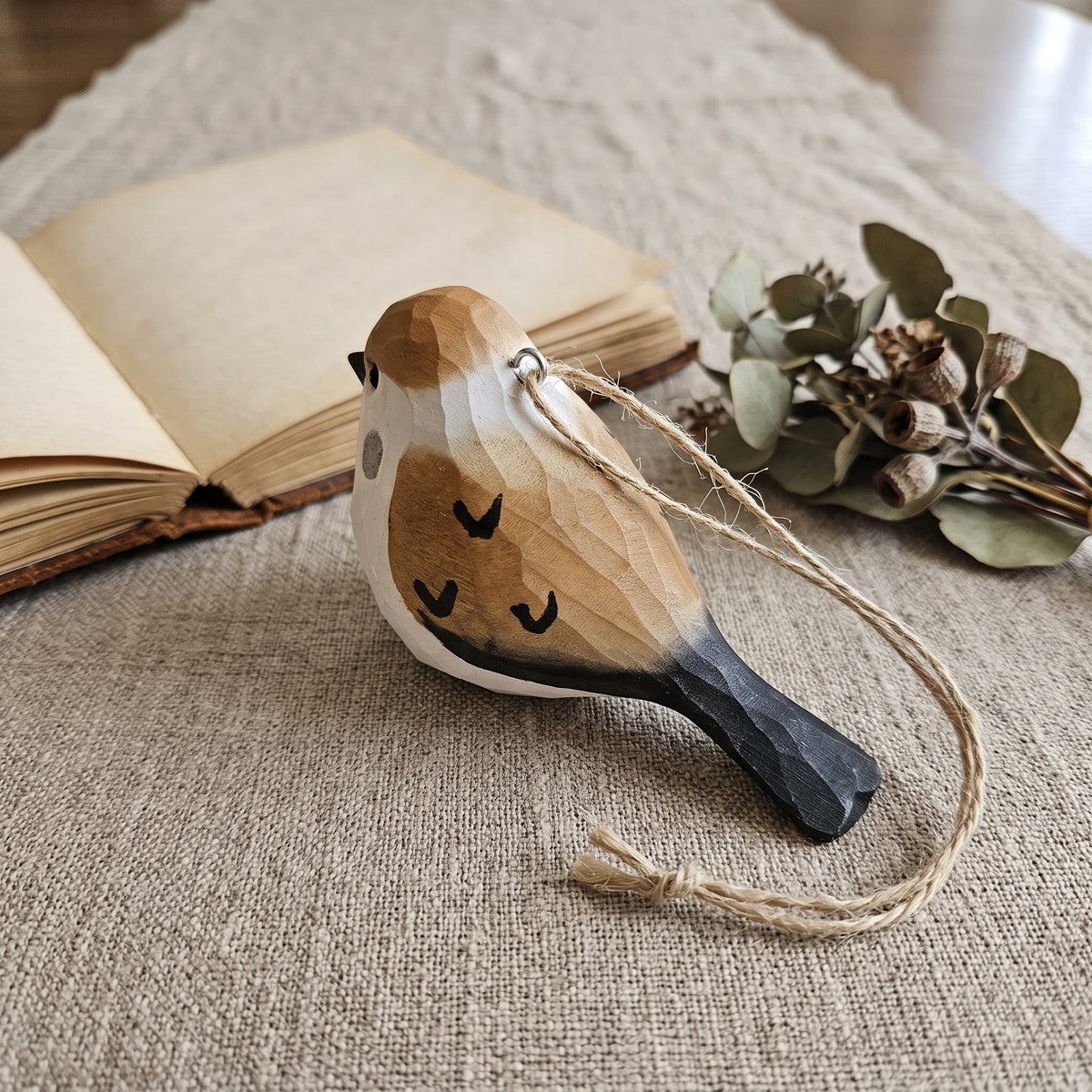 Hand-carved basswood sparrow ornament with jute string lying on a linen runner beside an open book