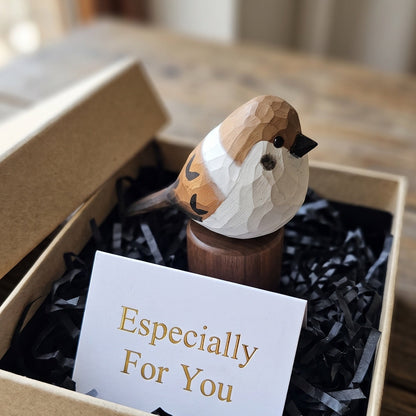 A small carved sparrow figurine perched on a wooden stand, presented inside a kraft gift box with black shredded paper filler and a card that reads 'Especially For You.' The box sits on a wooden table in a softly blurred indoor setting.