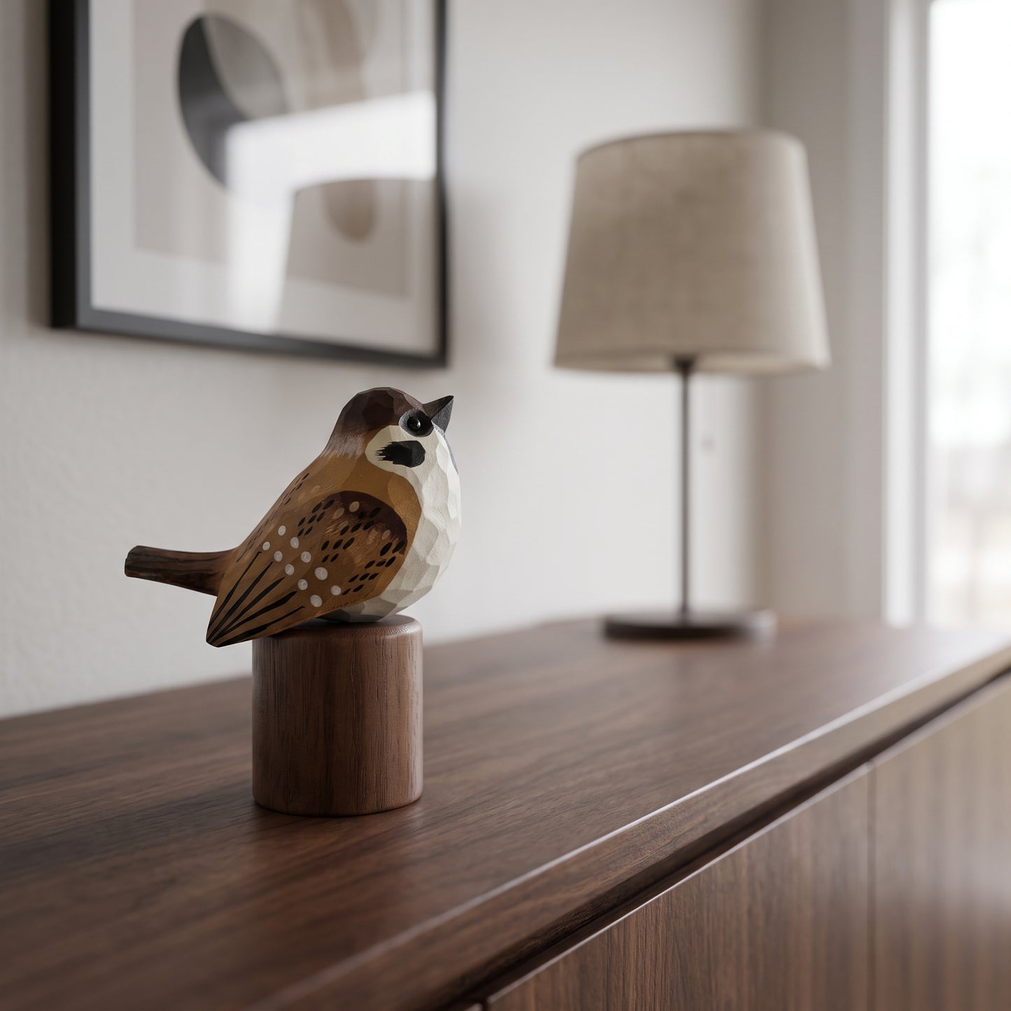 A small geometric Sparrow figurine in brown, beige, and black sits on a cylindrical wooden base on a dark wood console table, with a blurred lamp and framed wall art in the background.