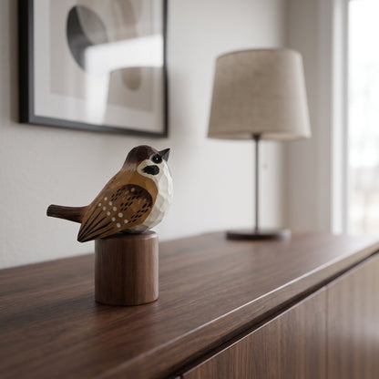 A small geometric Sparrow figurine in brown, beige, and black sits on a cylindrical wooden base on a dark wood console table, with a blurred lamp and framed wall art in the background.
