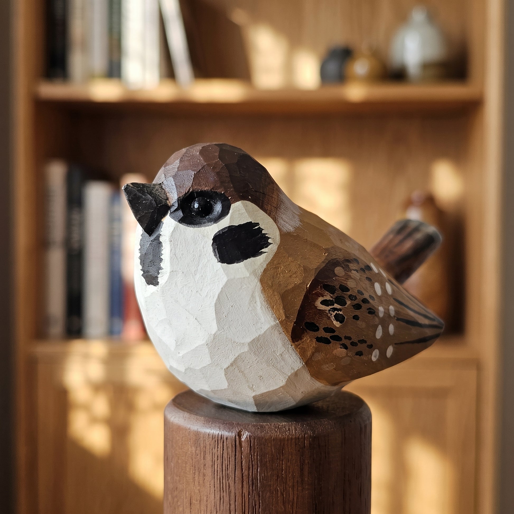 A hand-carved wooden Sparrow sculpture with a geometric, faceted finish, perched on a wooden post in front of a softly blurred wooden bookshelf.