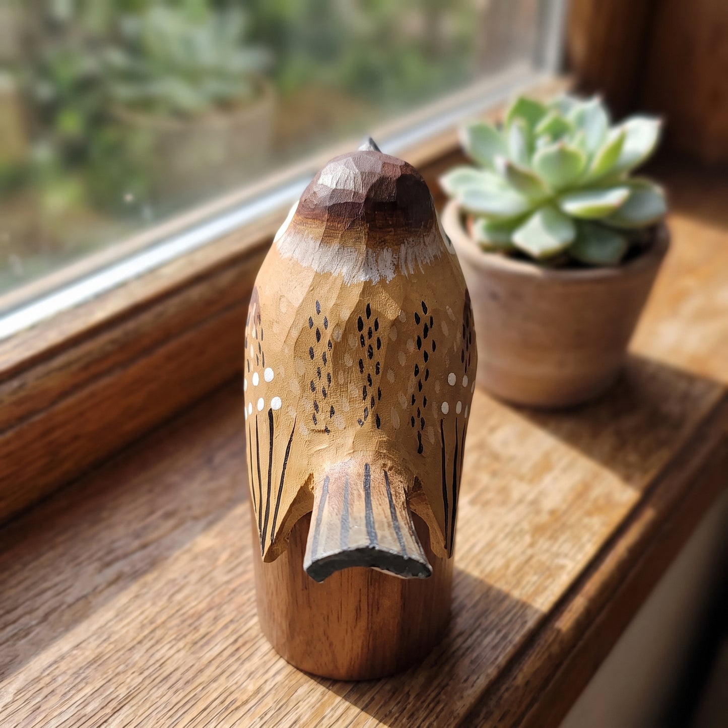 A hand-carved wooden Sparrow figurine with intricate geometric patterns and painted details, displayed on a wooden windowsill next to a small potted succulent.
