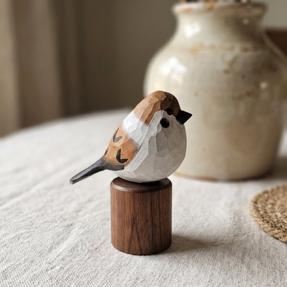 A hand-carved wooden Sparrow  figurine with a faceted geometric pattern, perched on a small cylindrical wooden base. The bird features natural wood tones of brown, beige, and white, with a small black beak.